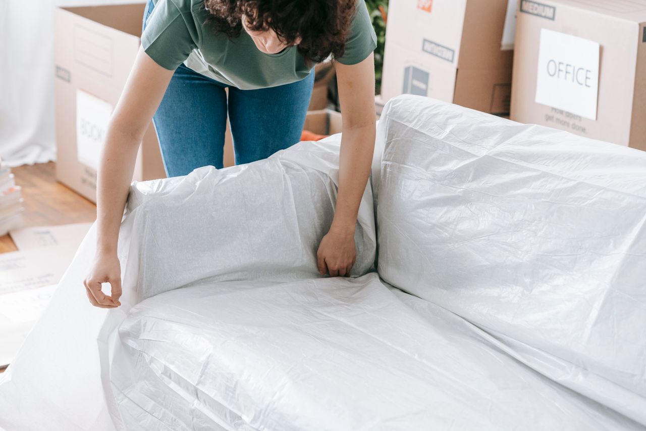 heros-img Une femme emballe soigneusement un canapé dans un matériau de protection tout en préparant des cartons dans une pièce lumineuse.
